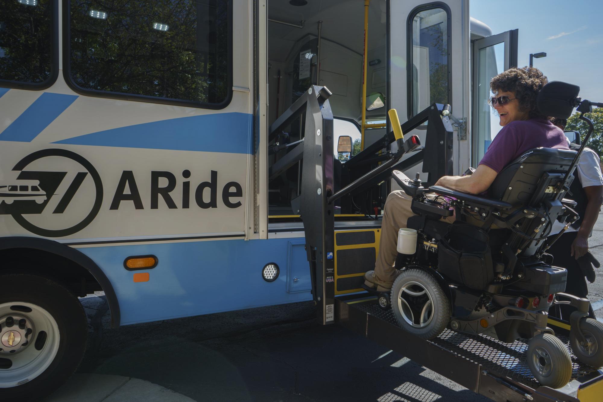 A passenger in a wheelchair is lifted up to ride on the A-Ride bus.