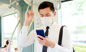 Male with face covering looking at phone while riding the bus and holding onto hand strap